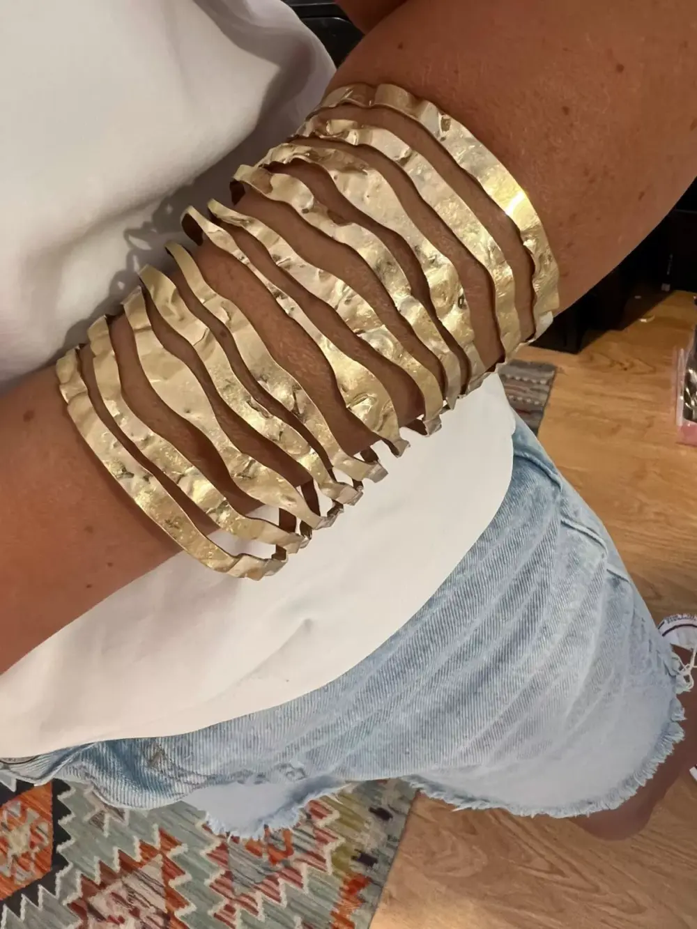 Braçadeira larga em latão com banho dourado, destacando-se pelo seu design ondulado e textura martelada que reflete a luz com elegância. Uma peça moderna e imponente que transforma qualquer look
